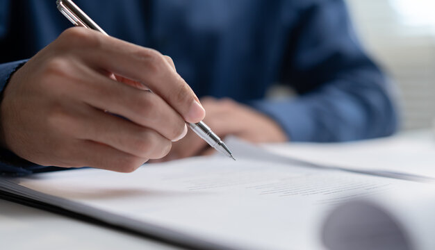 Close-up of businessman hand holding pen for signing legal contract document at office desk. Male corporate executive writing financial report, business agreement and deal concept.