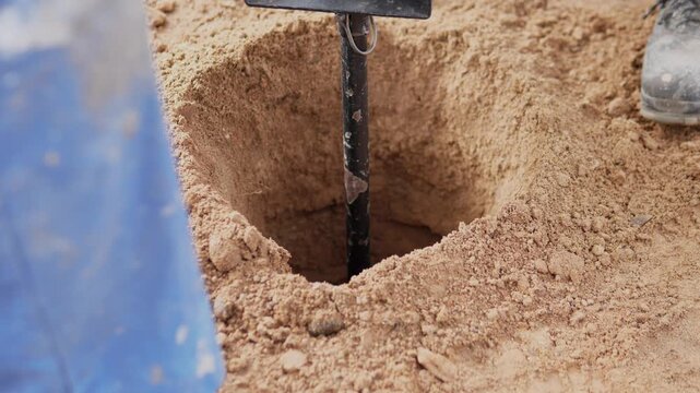 Tight elevated view of deep square hole in sandy soil with black digging tool standing upright, blue blur at left edge, and worker shoe at upper right, under even natural daylight with warm beige