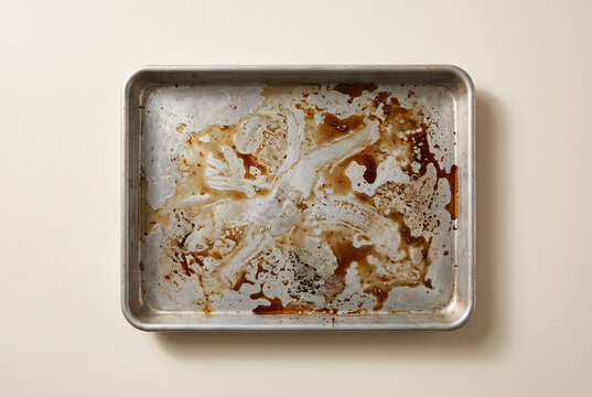 Top view of a dirty greasy baking sheet on a beige background. Unwashed aluminum metal pan covered in burnt food residue and oil. Flat lay of messy kitchenware