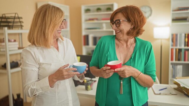 Woman holding coffee cup and smiling while talking with a colleague inside an office building; friendliness casual conversation.