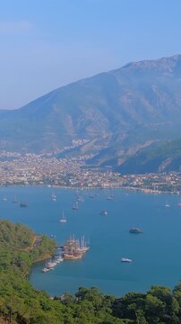 Vertical video: scenic view of Fethiye Bay with anchored and moving sailing boats, gulets under the clear blue sky. The bay is surrounded by lush green hills and distant mountains