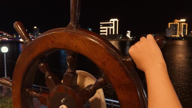 Wooden ship wheel on a han river cruise boat with hands gently turning it, navigating through the water at night, showcasing the vibrant city lights of da nang in vietnam