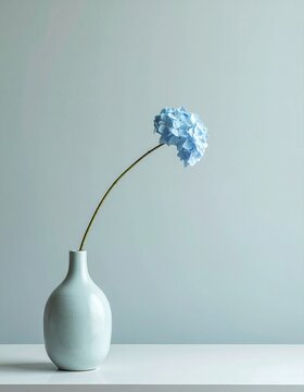 A single blue hydrangea flower with a curved stem in a minimalist ceramic vase on a white table against a soft gray background.