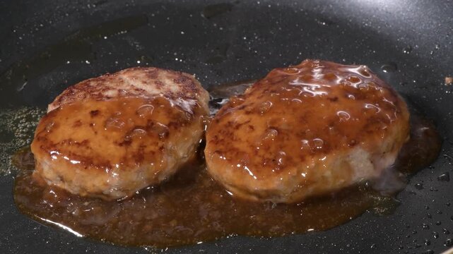 Two hamburger steak patties simmering in glossy brown sauce on a black frying pan
