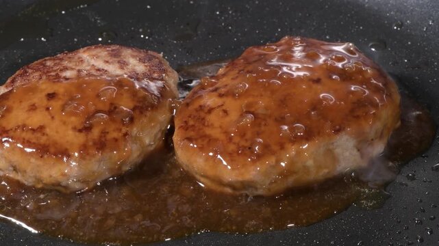 Two hamburger steak patties simmering in glossy brown sauce on a black frying pan