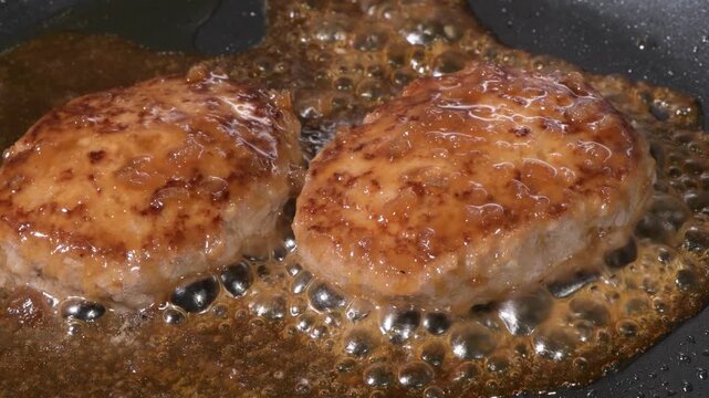Two hamburger steak patties simmering in glossy brown sauce on a black frying pan