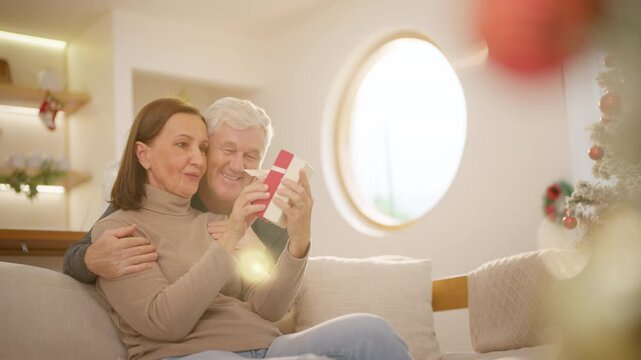 Family celebrating Christmas at home. Senior husband gives gift to wife while relaxing together in living room. Happy Caucasian older couple sharing love, surprise and festive holiday tradition.