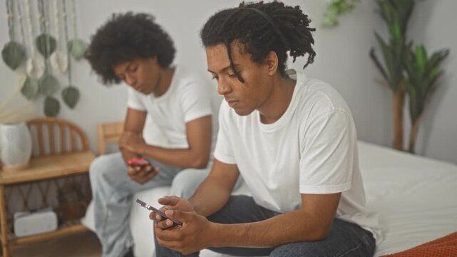 Man sitting on bed holding smartphone and smiling while friend in background scrolls phone in a bedroom inside a building; relaxed friendship.