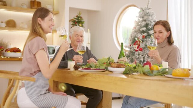 Senior parents and adult daughter enjoy Christmas dinner together at home. Happy family sharing festive holiday meal with roast chicken, salad and wine around dining table during seasonal celebration.