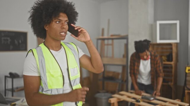 Man wearing yellow hi vis vest holds smartphone to ear while partner sands a wooden pallet in a building; concentration teamwork collaboration.