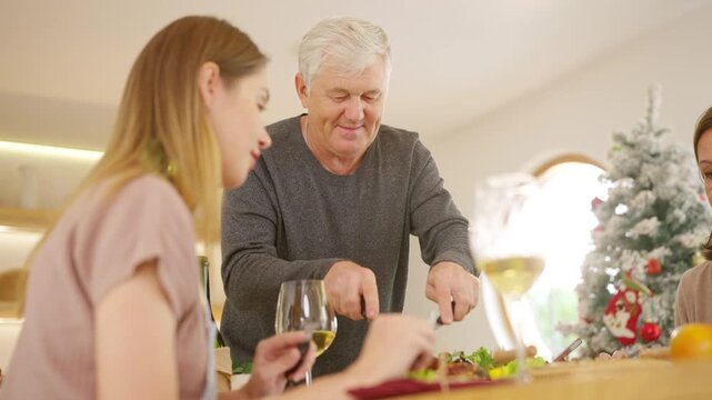 Senior parents and adult daughter enjoy Christmas dinner together at home. Happy family sharing festive holiday meal with roast chicken, salad and wine around dining table during seasonal celebration.