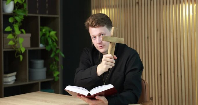 Priest in cassock with wooden cross reading Holy Bible at table indoors