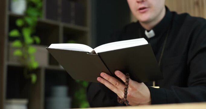 Priest in cassock with rosary reading Holy Bible indoors, closeup