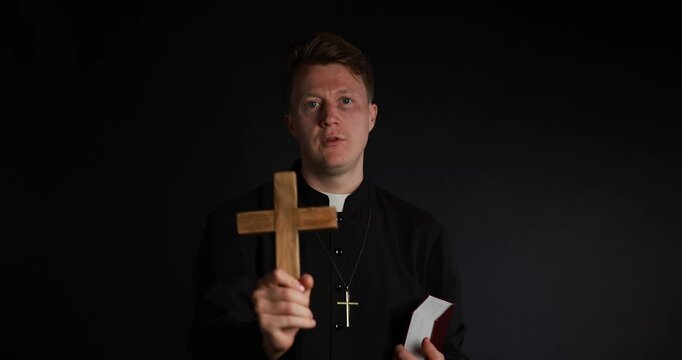 Priest in cassock with wooden cross and Holy Bible blessing on black background
