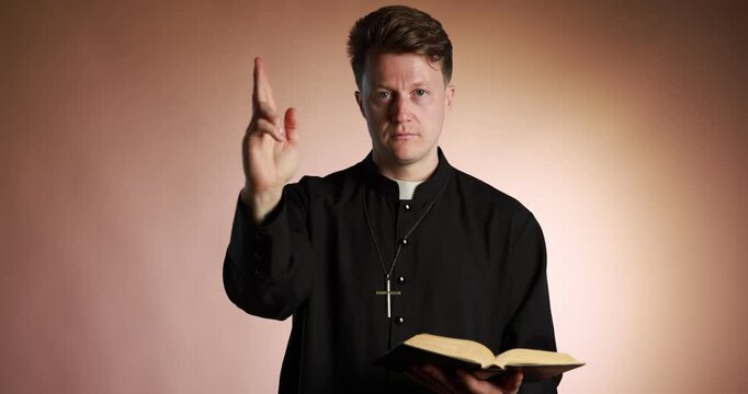 Priest in cassock with Holy Bible making blessing gesture on pale brown background