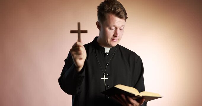 Priest in cassock with wooden cross reading Holy Bible on pale brown background