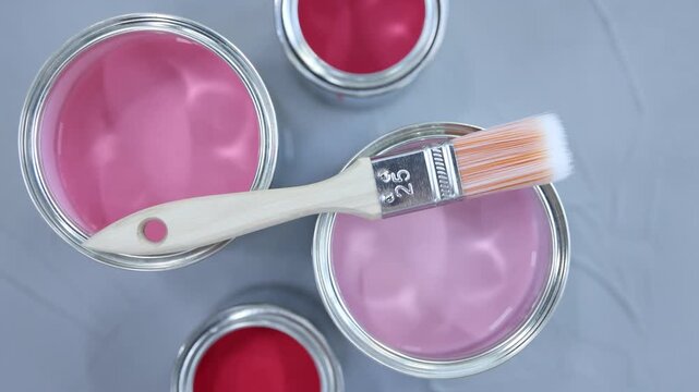 Cans with paint in shades of pink and brush on grey background, flat lay. Camera moving in