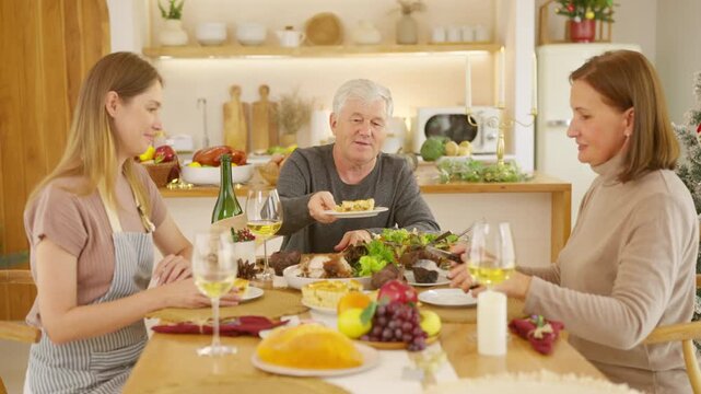 Senior parents and adult daughter enjoy Christmas dinner together at home. Happy family sharing festive holiday meal with roast chicken, salad and wine around dining table during seasonal celebration.