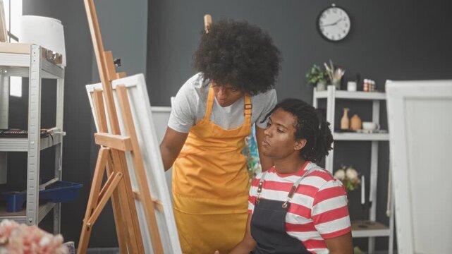 Two painters, both men, in aprons lean over a canvas, one pointing a paintbrush while the seated man holds a palette in an art studio; mentorship focus.