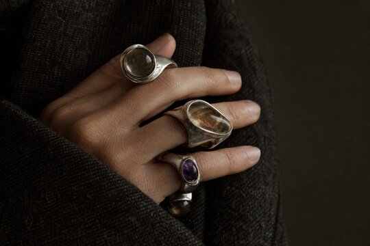 Close-up of elegant hand adorned with three distinctive silver rings featuring gemstone and textured designs, moody dark background, luxury jewelry fashion, minimalist portrait detail