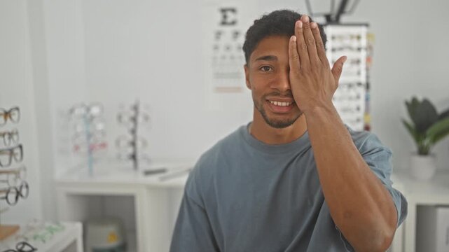 Man covering one eye with hand for an eye test in an optometry building with eyeglass racks and eye chart visible, smiling while seated; confidence vision care.