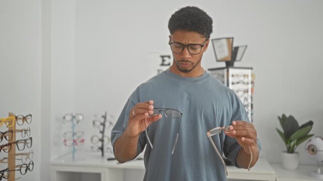 Young african american man holding two pairs of glasses while comparing frames and lenses at an optician store counter; confidence choice.