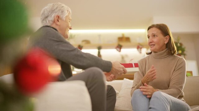 Family celebrating Christmas at home. Senior husband gives gift to wife while relaxing together in living room. Happy Caucasian older couple sharing love, surprise and festive holiday tradition.