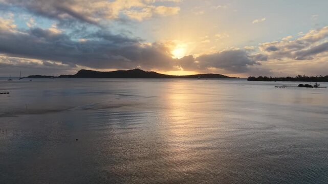 Aerial drone shot moving forward over shallow coastal waters, docked small motorboats, and the sun breaking through clouds during a golden sunset in Bora Bora