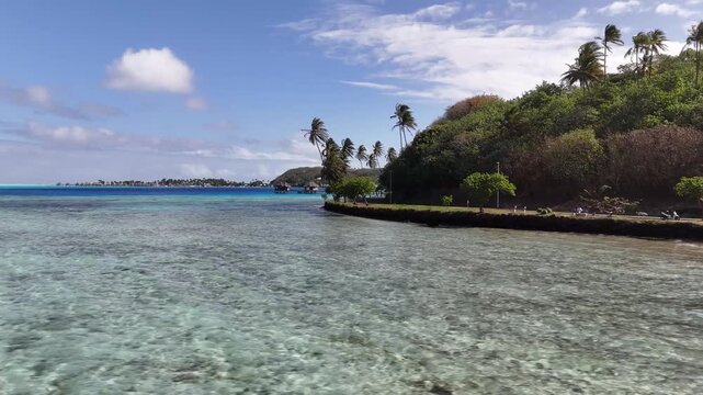 Aerial ateral tracking shot shifting left over a shallow turquoise lagoon, showcasing a paved coastal loop road, palm trees, and lush green hillsides in Bora Bora