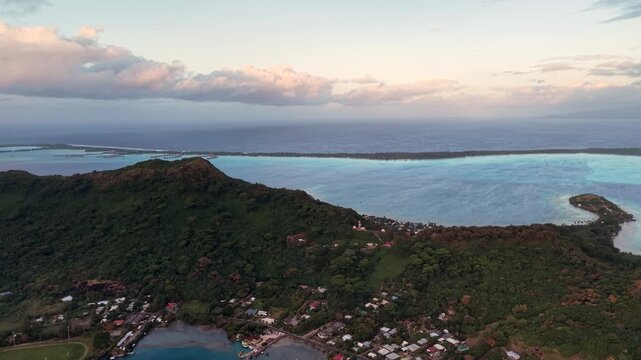 Aerial drone shot moving forward over a lush green ridge, revealing the turquoise coastal lagoon and overwater bungalows of Conrad Bora Bora Nui in Bora Bora