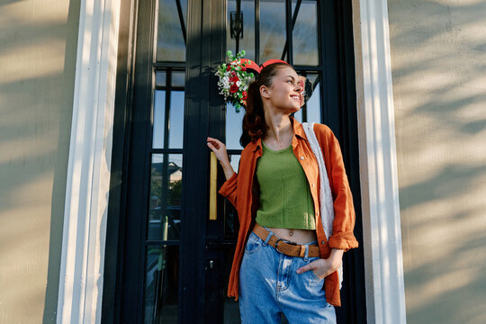 Smiling young woman in an orange shirt and jeans stands by a black door with a festive wreath, holding a bag. Concept of holidays and winter mood.