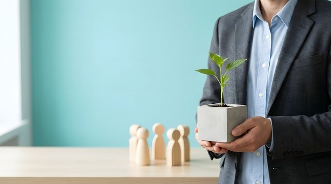 Person in a grey suit holding a small green plant in a square concrete pot, with wooden peg dolls standing on a light wooden table surface