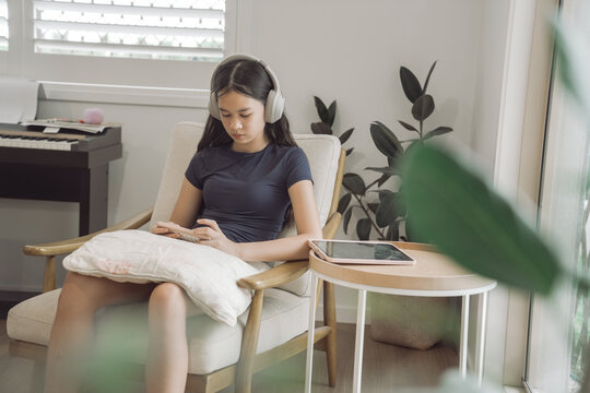 Teenager, headphones, smartphone, home, listening, technology, girl sitting in a comfortable armchair at home, using a mobile phone and wearing wireless headset in a modern living room.