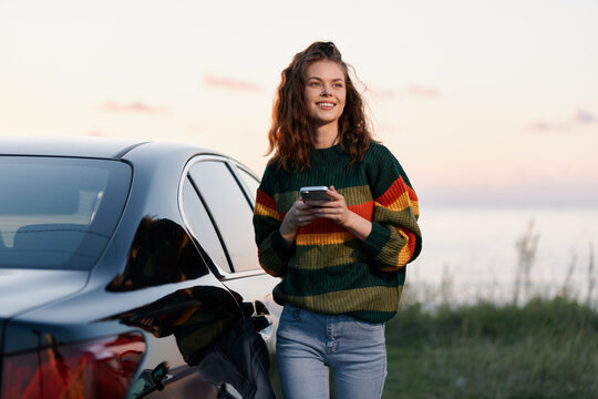Smiling young woman in colorful sweater stands by her car near the sea at sunset, using smartphone. Lifestyle concept of travel and communication.