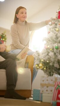 Senior couple decorating Christmas tree at home. Happy Caucasian older husband and wife enjoying festive holiday season together in living room with ornaments, gift boxes and seasonal celebration