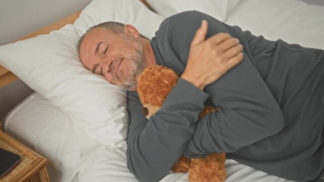 Middle aged man hugging teddy bear on bed in building, bearded and smiling, eyes closed with arm across chest; comfort nostalgia.