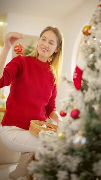 Adult woman decorating Christmas tree with ornaments at home. Happy Caucasian woman enjoying festive holiday preparation, seasonal family tradition and Christmas celebration in living room