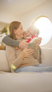 Family celebrating Christmas at home. Senior husband gives gift to wife while relaxing together in living room. Happy Caucasian older couple sharing love, surprise and festive holiday tradition.