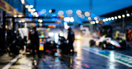 Obraz premium A blurred image of a racetrack during a nighttime Formula 1 event, featuring pit crews and cars under lights amidst rainy conditions.
