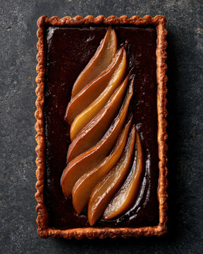 Rectangular dark chocolate tart topped with glossy poached pear slices, photographed overhead on a dramatic charcoal stone surface.