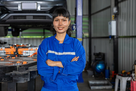 Confident female mechanic in blue coverall standing with crossed arms inside automotive garage beside electric vehicle assembly area