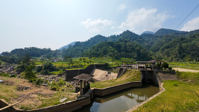 Wide angle view of a concrete river dam and irrigation canal surrounded by green rice fields and lush mountains in West Java, Indonesia