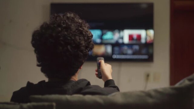 Person with curly hair zapping through streaming channels on smart TV with remote control at home