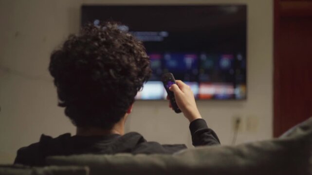 Young adult zapping through streaming platform on smart TV with remote control at night