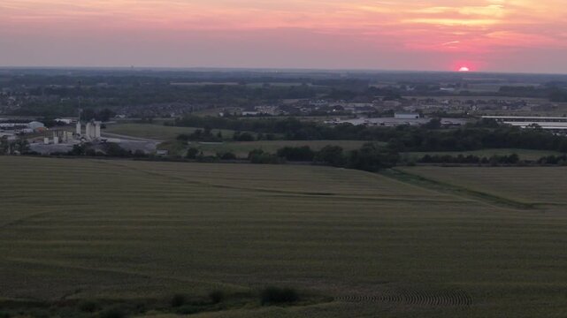 Aerial drone view vast Midwestern cornfield at sunset bordering suburban residential development and industrial infrastructure in Kansas