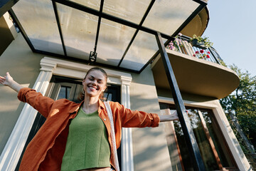 A joyful young woman in an orange jacket and green top stands with open arms under a glass canopy near her house, smiling happily, celebrating freedom and a new home concept. © SHOTPRIME STUDIO