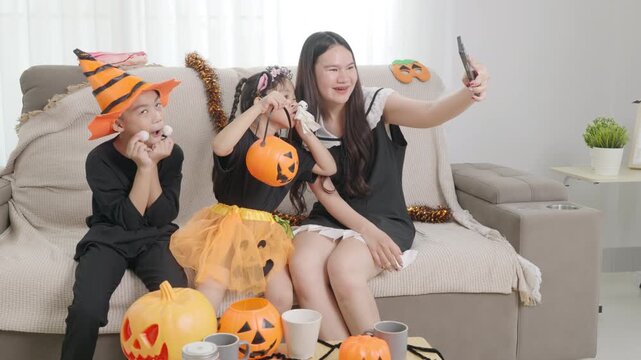 Asian family, woman, boy, girl celebrating festive halloween holiday at home, taking joyful selfie with smartphone, enjoying fun costumes, pumpkins, holiday decorations.