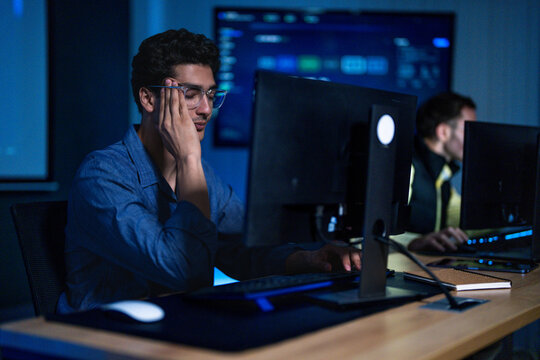 A male technology worker resting his head in one hand at his desk sighs quietly as another alert appears on the display at a developer desk washed in blue light from surrounding displays