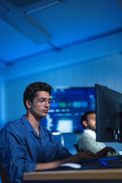 A tired male analyst rubbing his face in front of glowing monitors presses a palm against his forehead while the screens keep updating inside a modern digital office