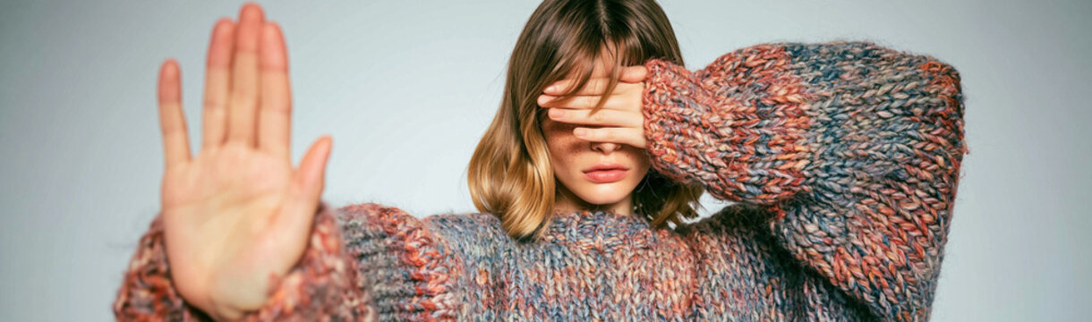 Stop, Don't Look: A young woman with her hand raised in a stop gesture and eyes covered, conveying a message of refusal or discomfort, her patterned sweater adding a textural contrast.